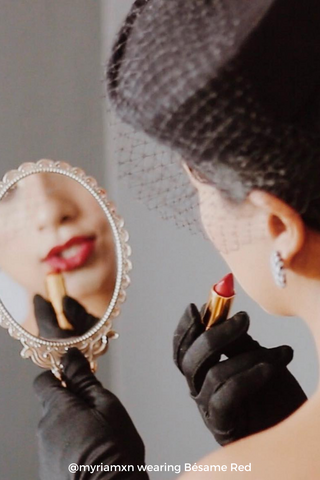 Woman wearing black gloves and black hat holding a mirror and applying Bésame's Red Lipstick
