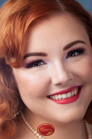 Red haired woman smiling wearing a perl necklace and Bésame's red lipstick